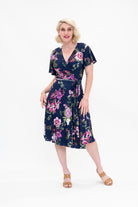 Model wearing the Hazel knee-length floral wrap dress with flutter sleeves by By Samantha Melbourne. Curve-friendly fit with a deep navy base and pink botanical print.