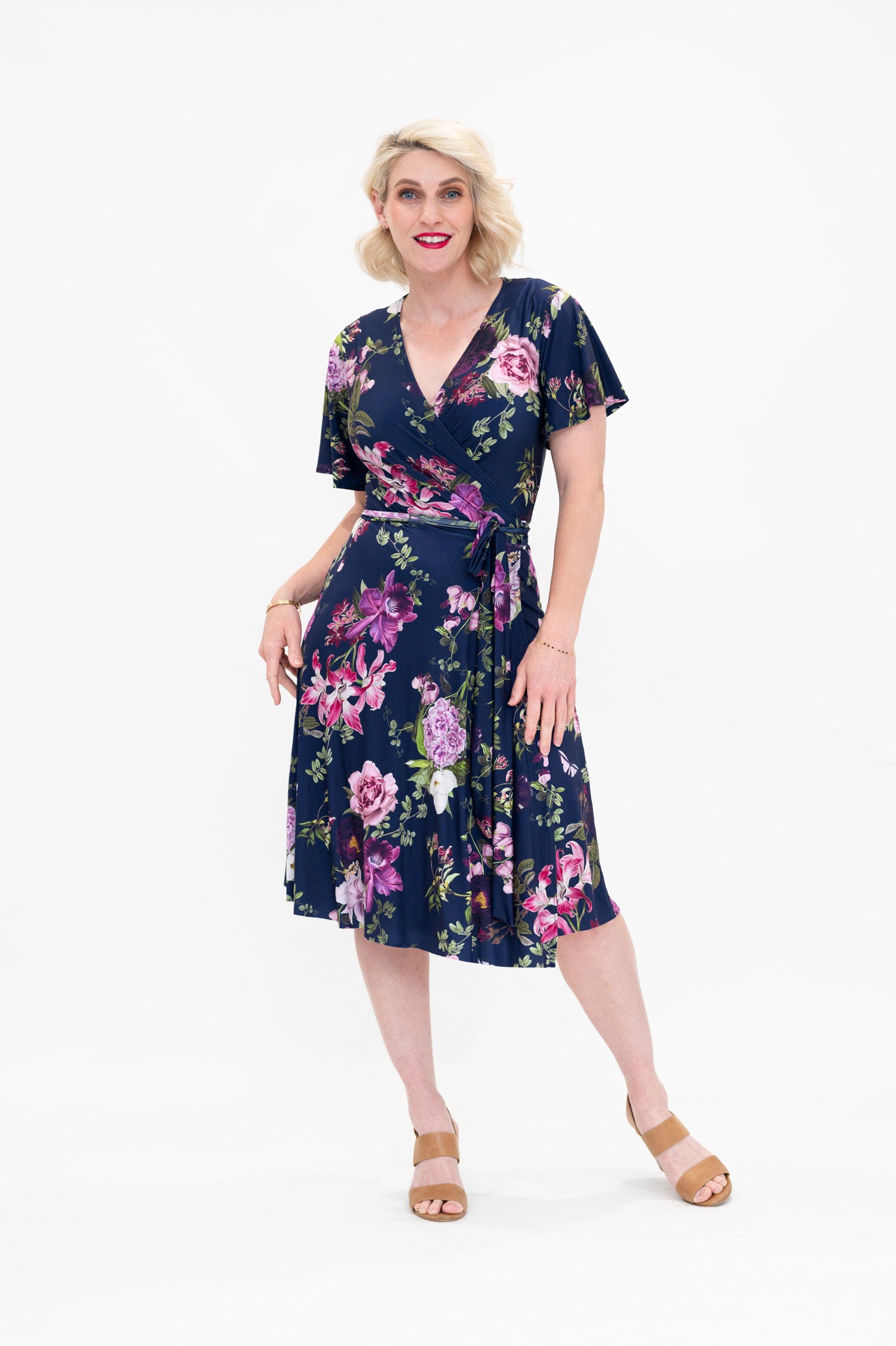 Model wearing the Hazel knee-length floral wrap dress with flutter sleeves by By Samantha Melbourne. Curve-friendly fit with a deep navy base and pink botanical print.