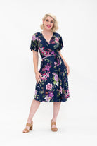 Model wearing the Hazel knee-length floral wrap dress with flutter sleeves by By Samantha Melbourne. Curve-friendly fit with a deep navy base and pink botanical print.