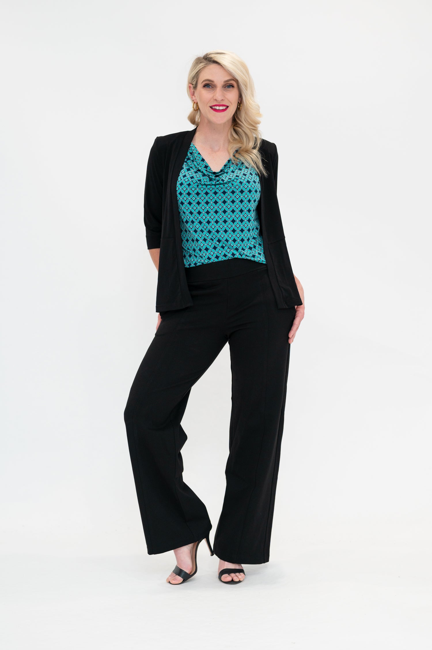 The female model wears a cowl neck top with 3/4 sleeves, the top is tucked into black pants. The print is small scale deep navy, white and teal geometric diamond shape pattern. The model wears the soft black jacket on the top of the cowl neck top