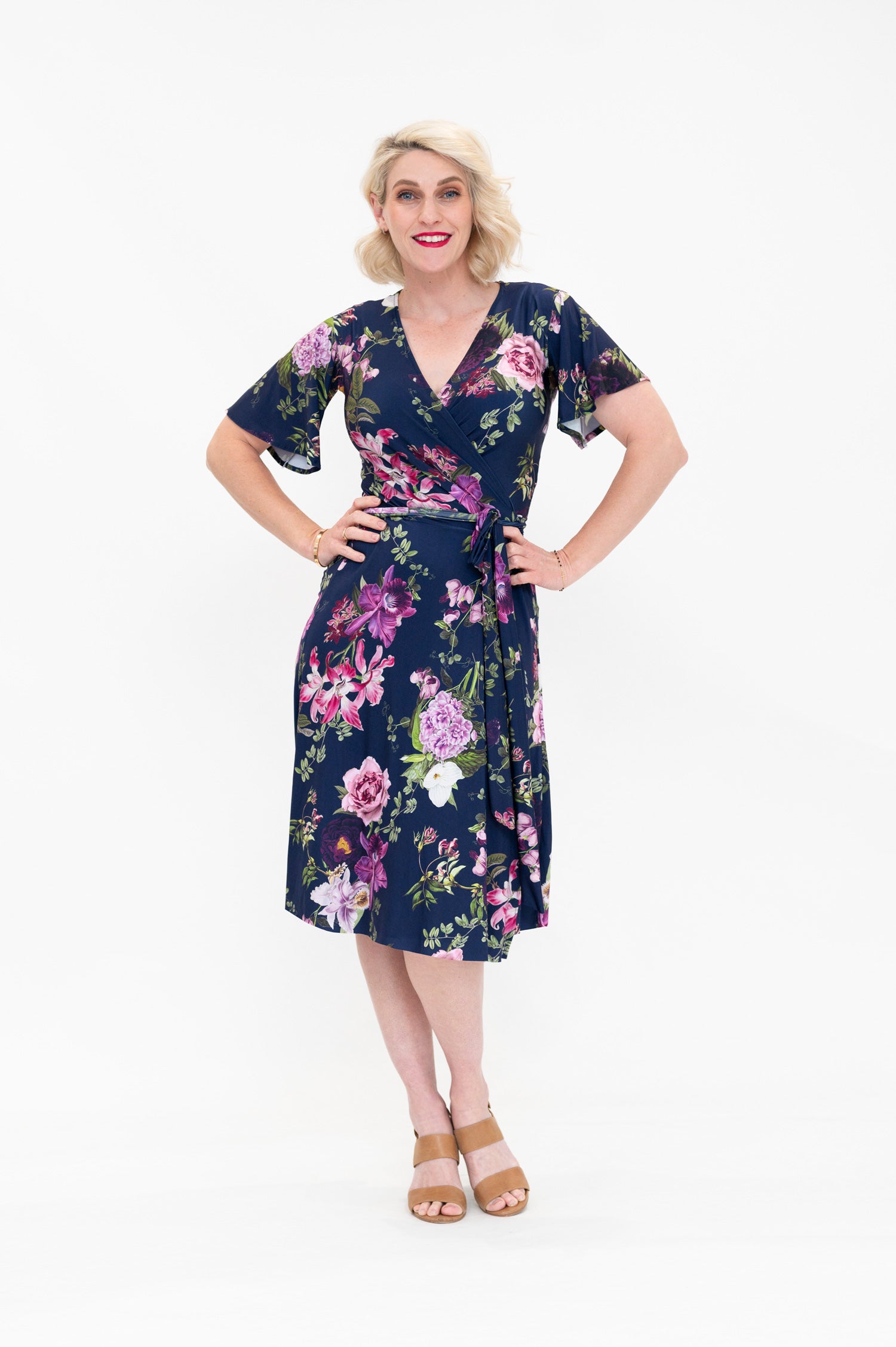 Model wearing the Hazel knee-length floral wrap dress with flutter sleeves by By Samantha Melbourne. Curve-friendly fit with a deep navy base and pink botanical print.