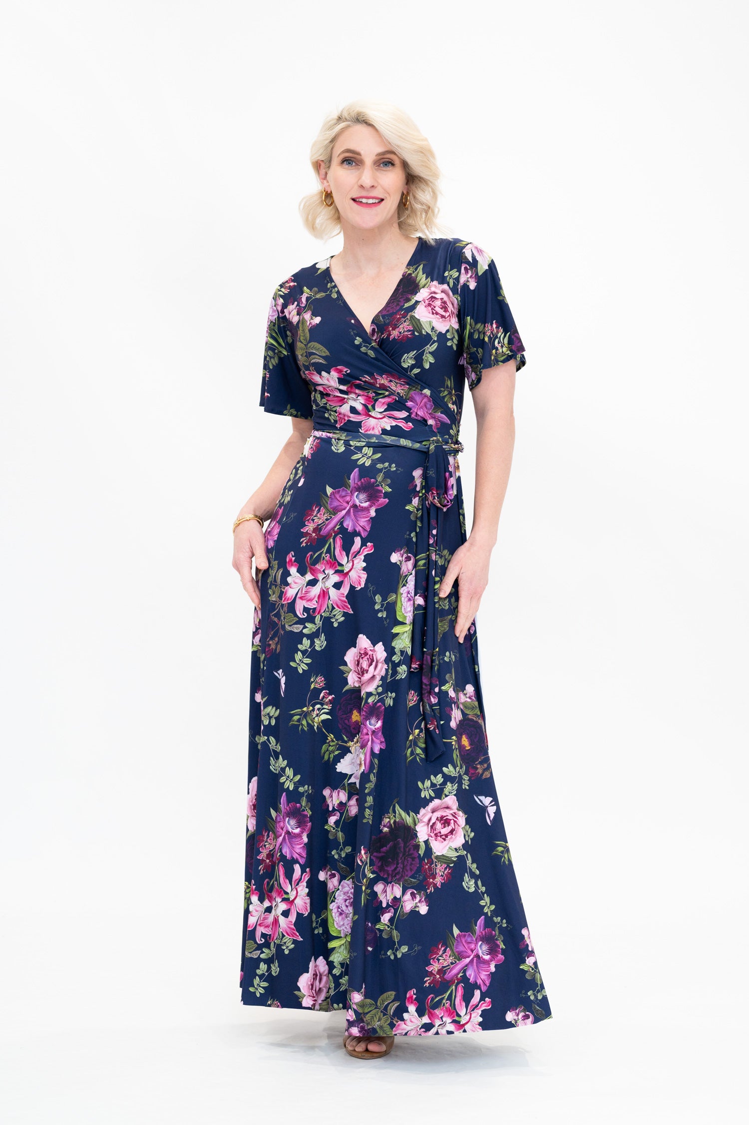 Model wearing the Hazel maxi floral wrap dress with flutter sleeves by By Samantha Melbourne. Curve-friendly fit with a deep navy base and pink botanical print.