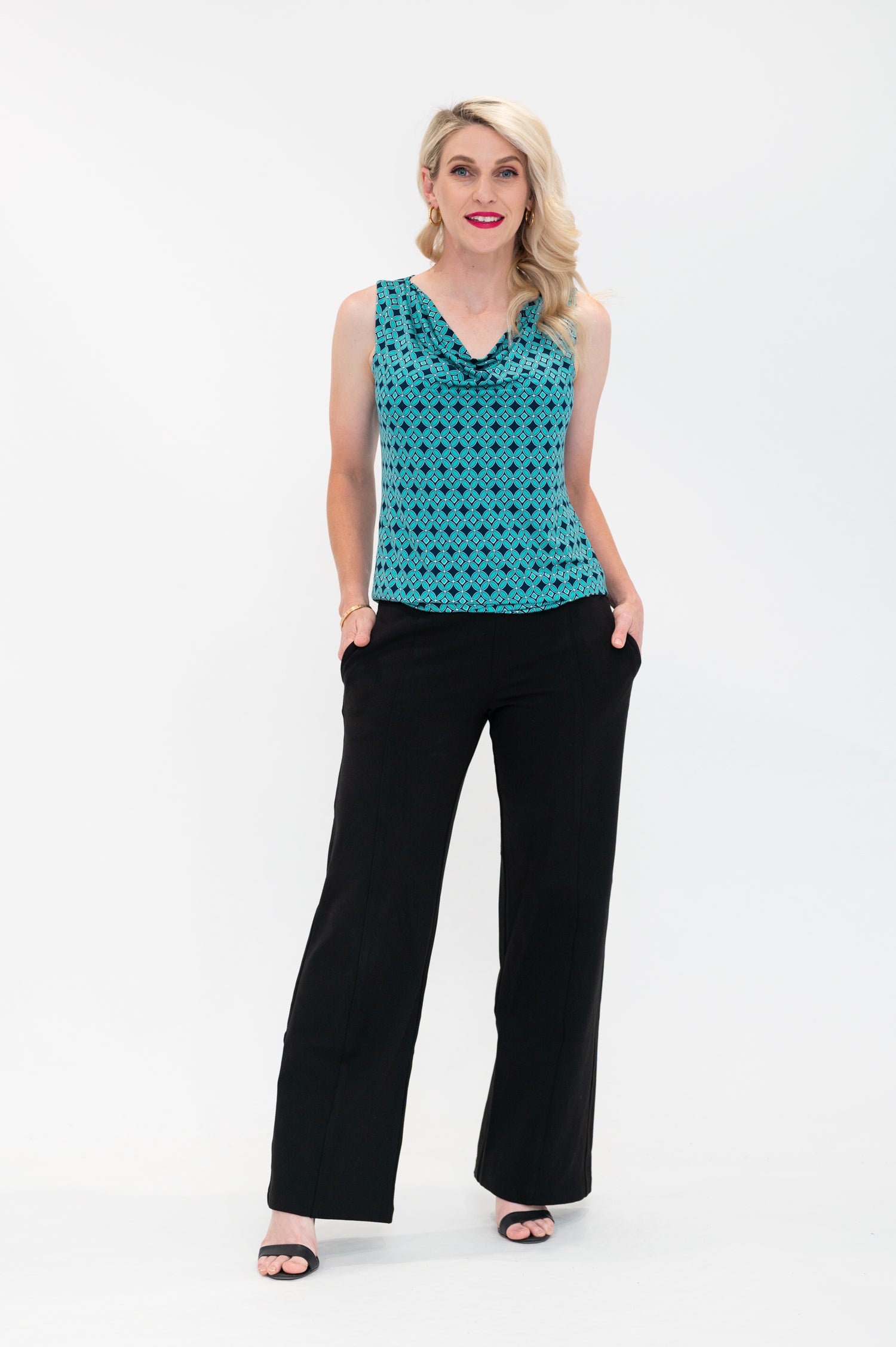 The female model wears a cowl neck top with no sleeves, the top is untucked in black pants. The print is small scale deep navy, white and teal geometric diamond shape pattern