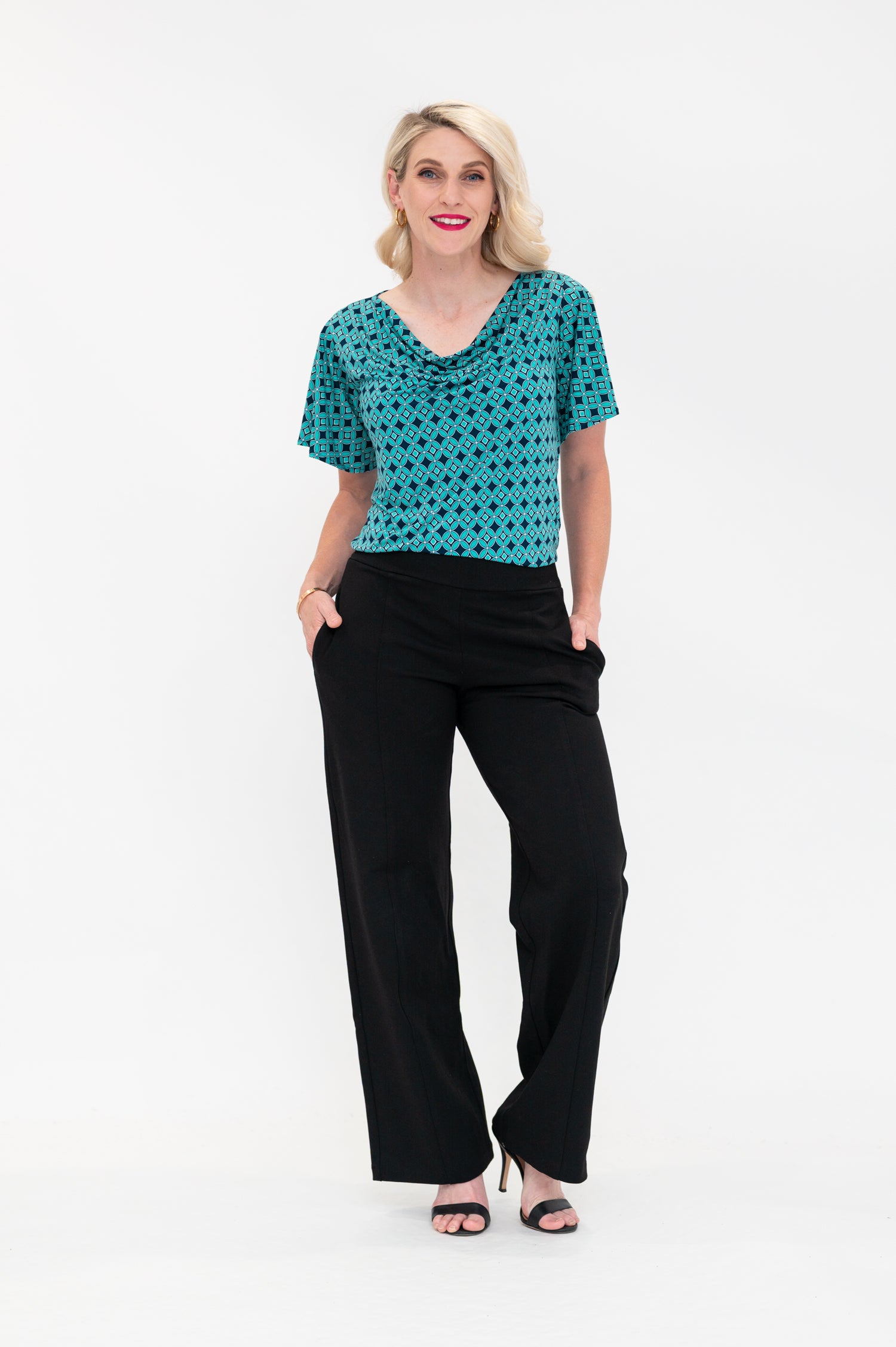 The female model wears a cowl neck top with flutter sleeves, the top is tucked into black pants. The print is small scale deep navy, white and teal geometric diamond shape pattern