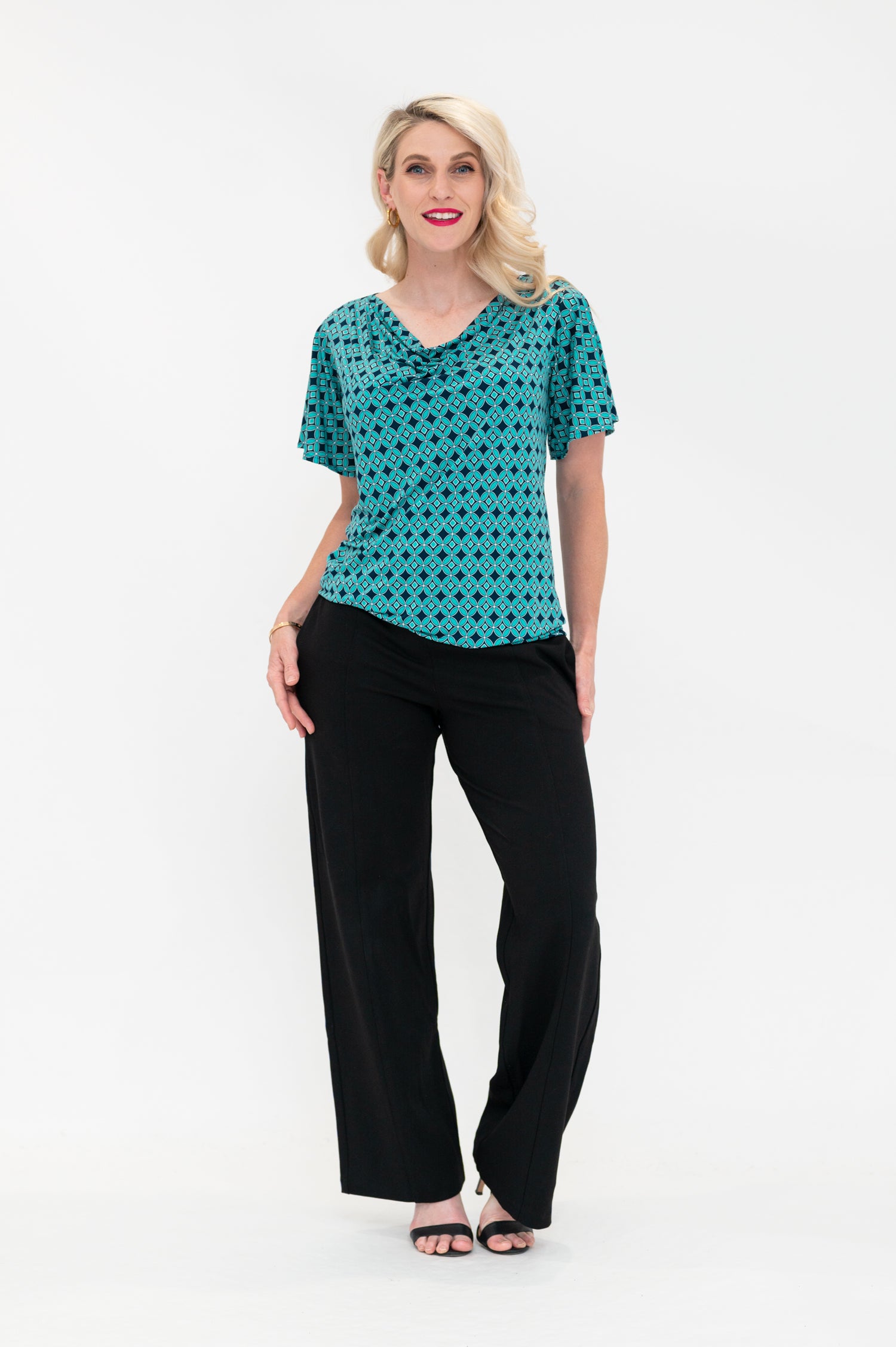 The female model wears a cowl neck top with flutter sleeves, the top is untucked in black pants. The print is small scale deep navy, white and teal geometric diamond shape pattern