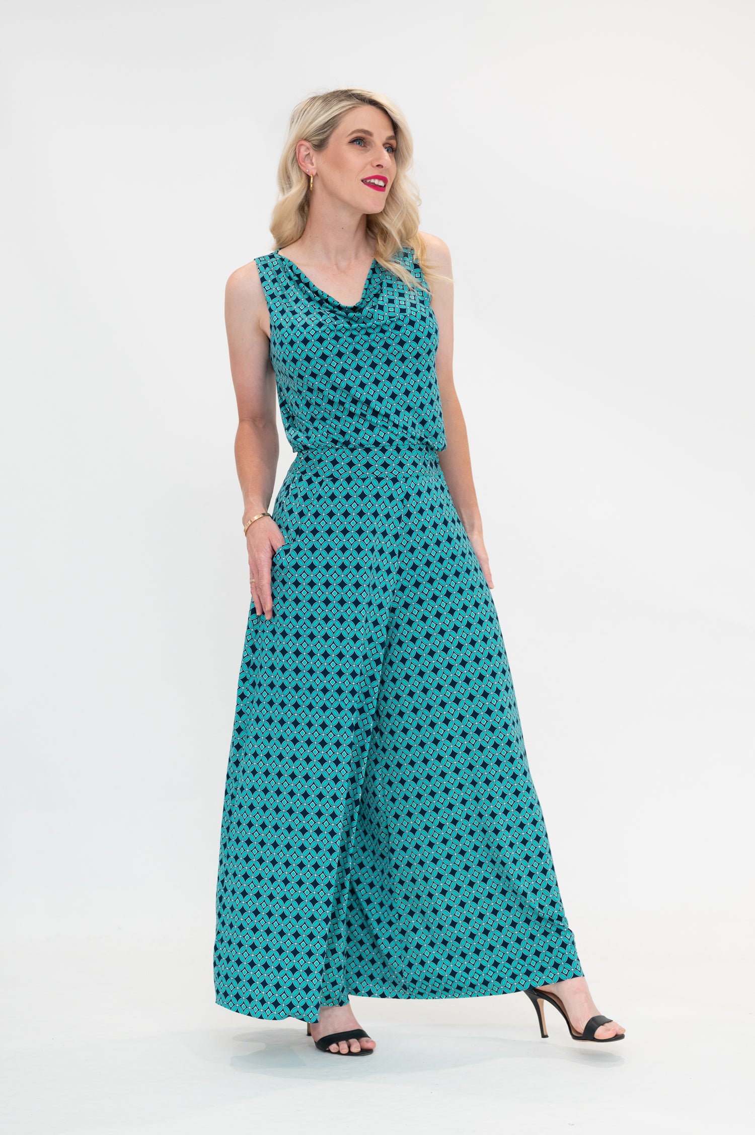 The female model wears a cowl neck top with no sleeves, the top is tucked in palazzo pants. The print is a small-scale deep navy, white and teal geometric diamond shape pattern. Side view
