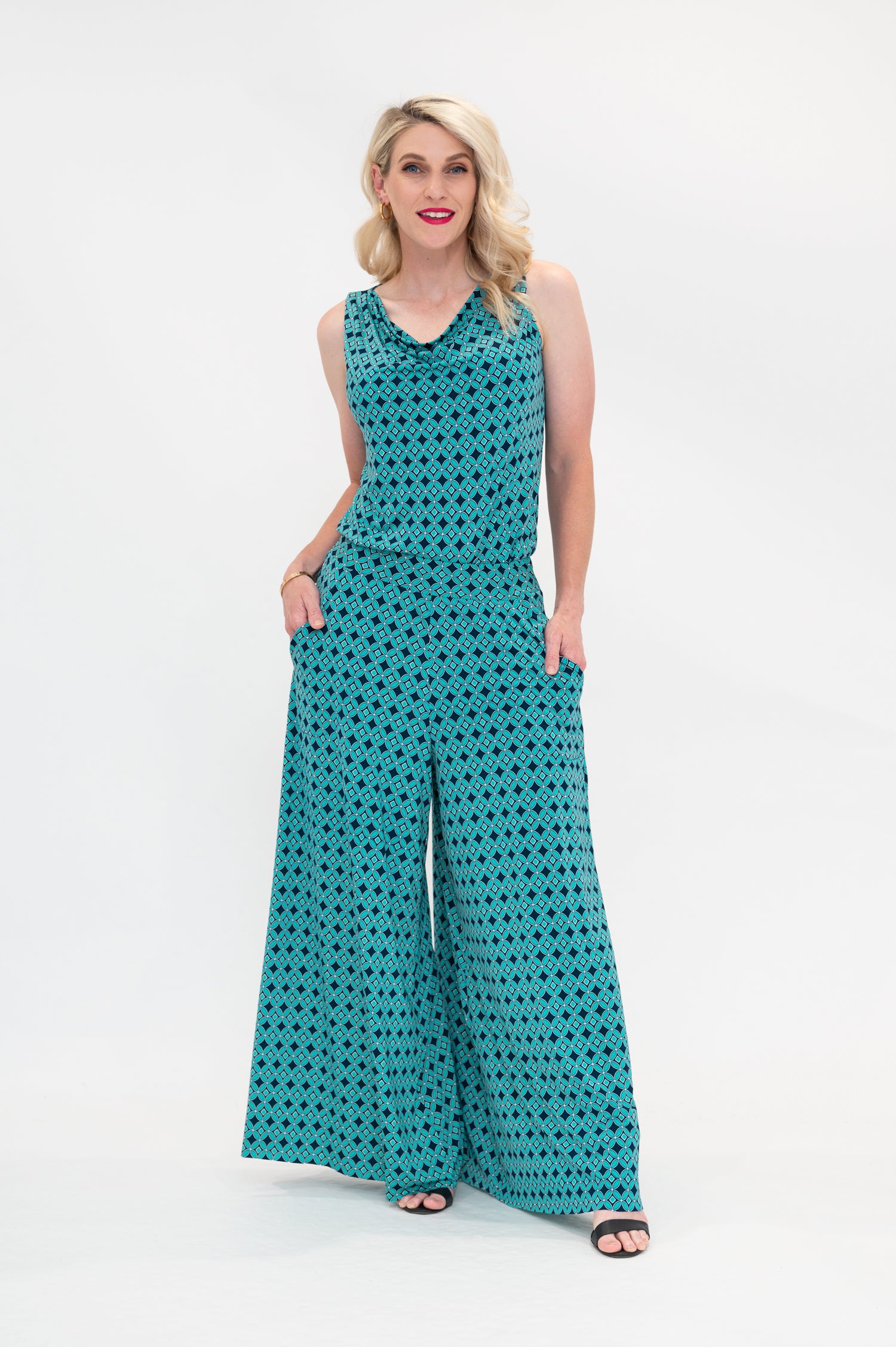 The female model wears a cowl neck top with no sleeves, the top is tucked in palazzo pants. The print is a small-scale deep navy, white and teal geometric diamond shape pattern. Front view