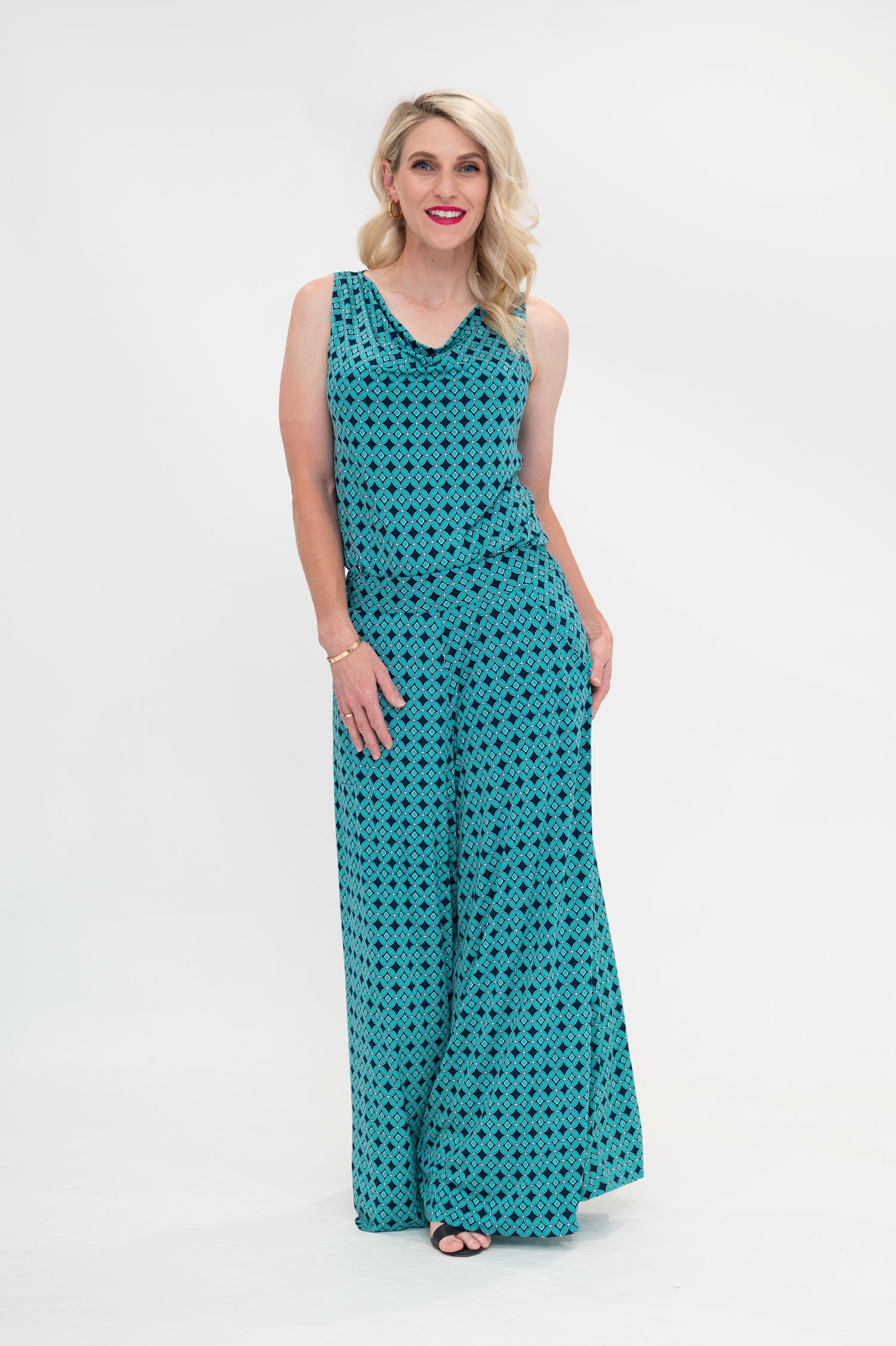 The female model wears a cowl neck top with no sleeves, the top is tucked in palazzo pants. The print is a small-scale deep navy, white and teal geometric diamond shape pattern. Front view