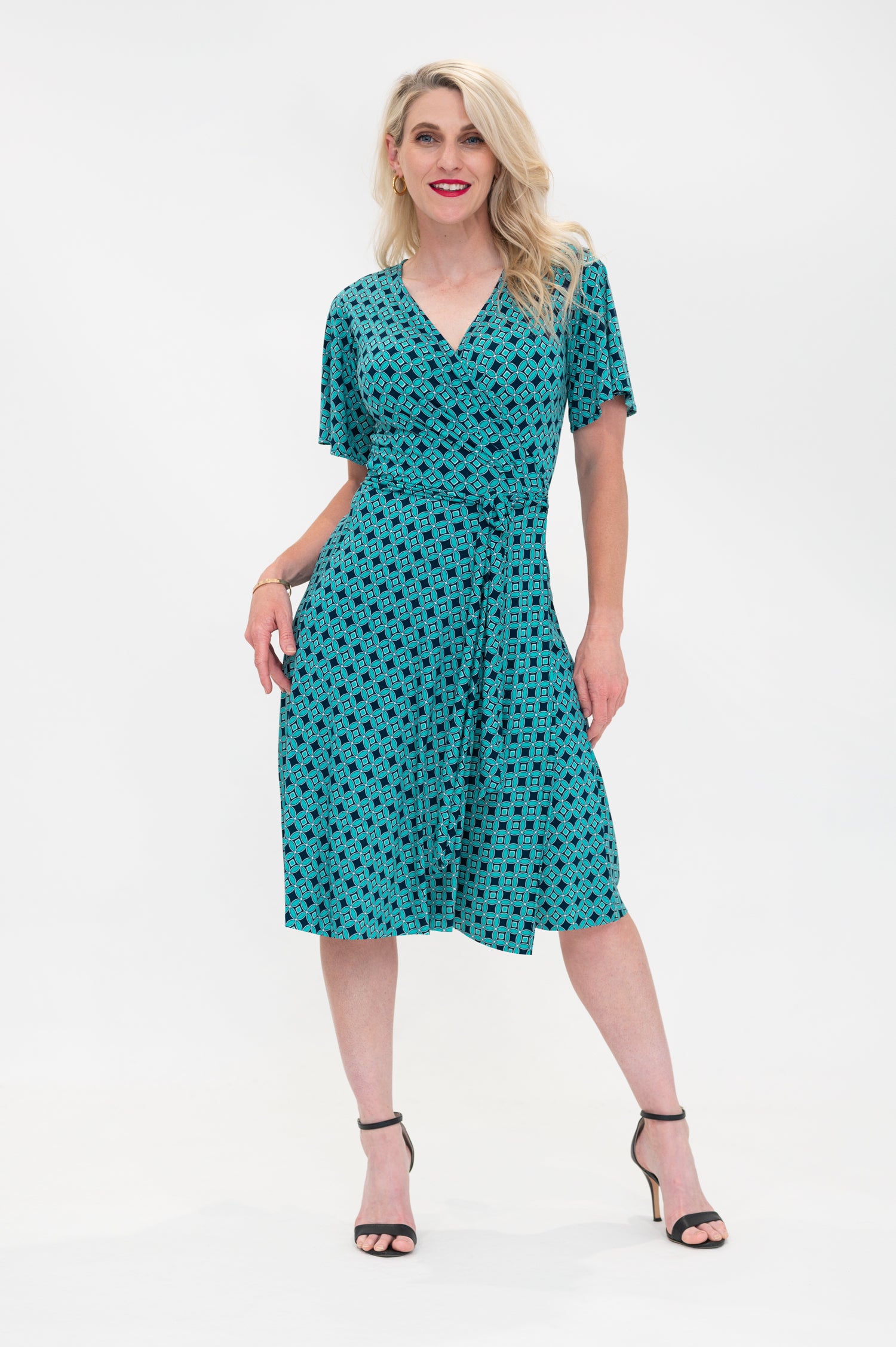 The female model wears a wrap dress in knee length with flutter sleeves. The print is a small-scale deep navy, white and teal geometric diamond shape pattern. Front view