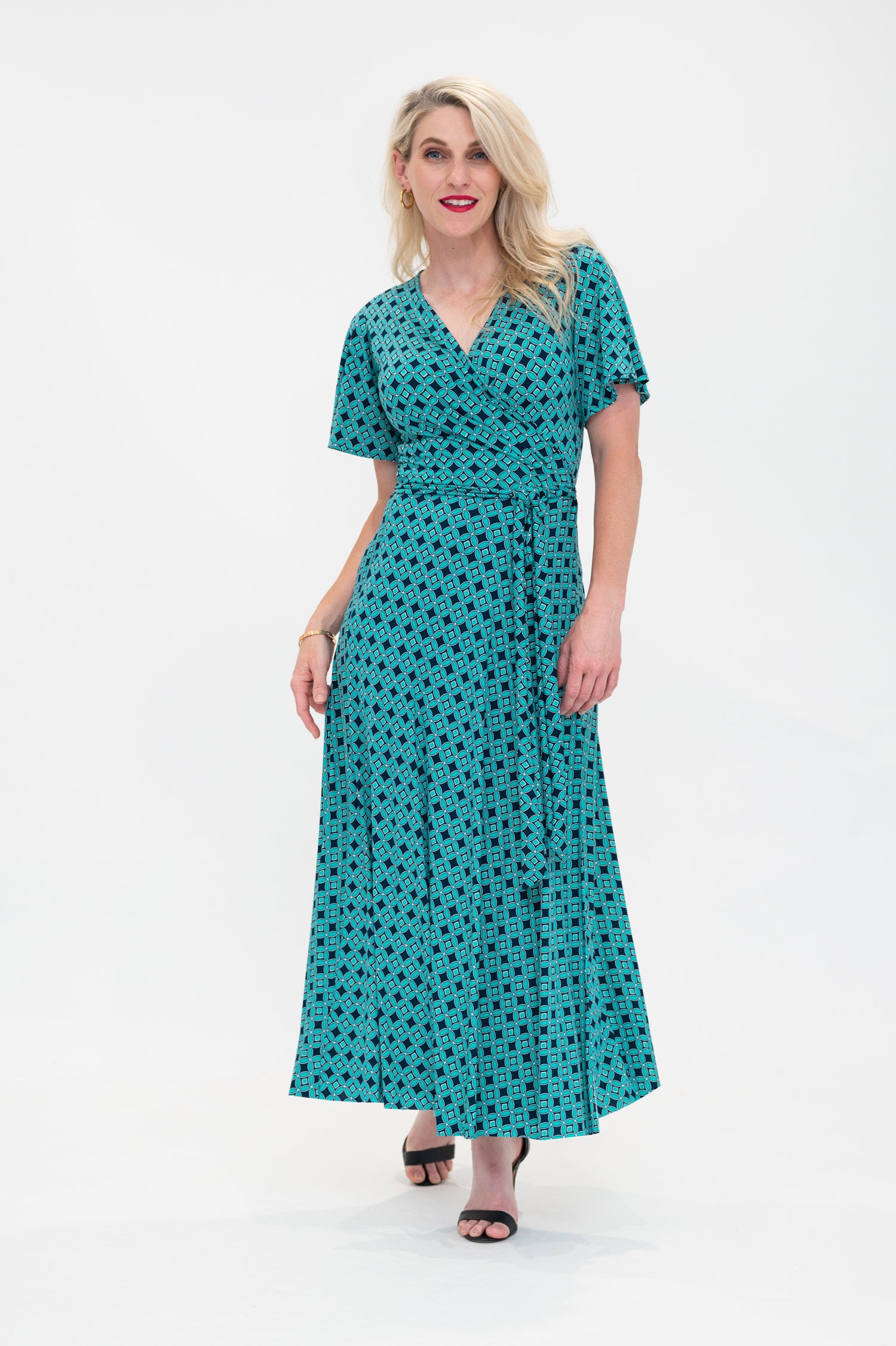 The female model wears a wrap dress in midi length (ankle length) with flutter sleeves. The print is a small-scale deep navy, white and teal geometric diamond shape pattern. Front view