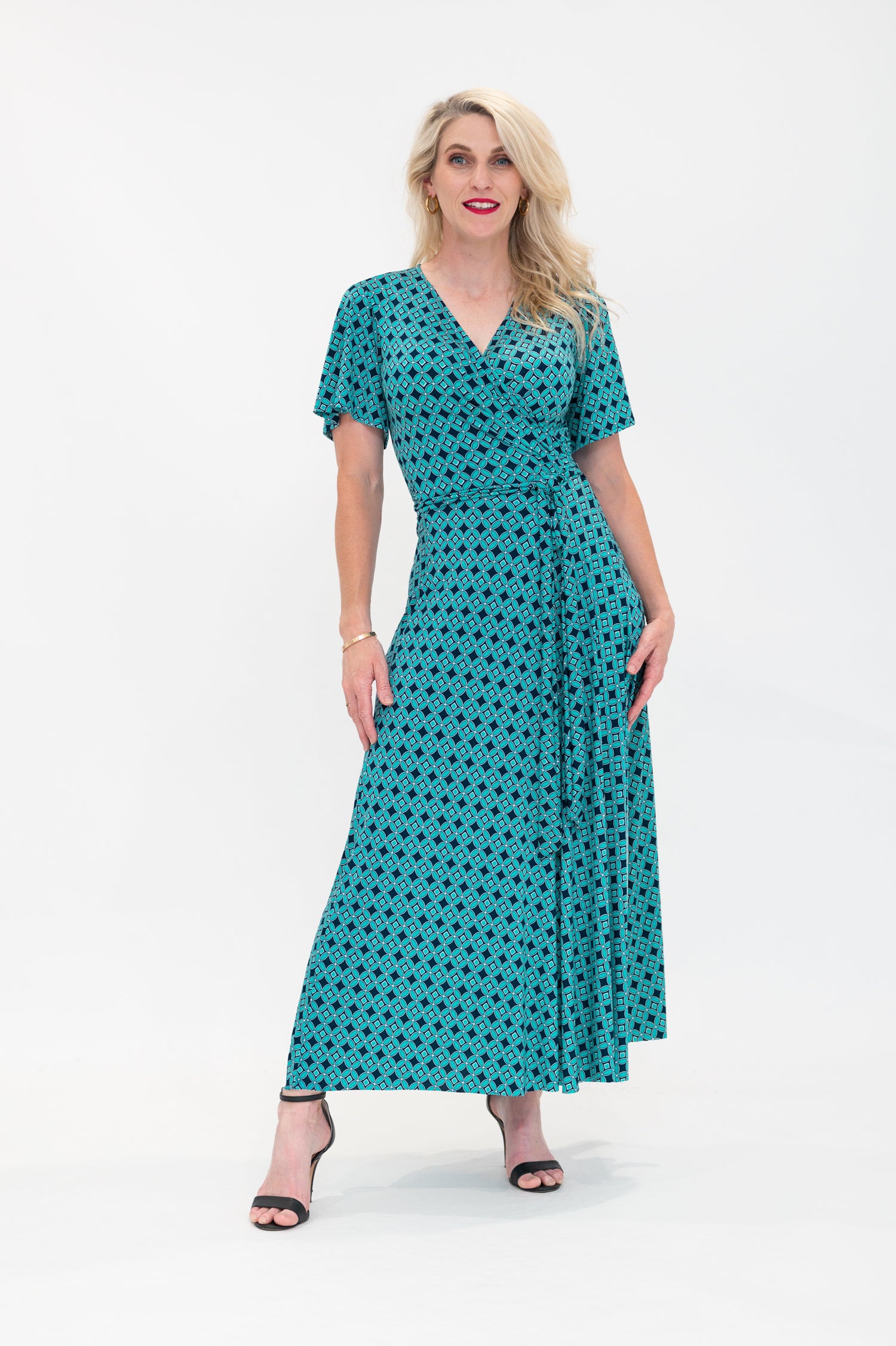 The female model wears a wrap dress in midi length (ankle length) with flutter sleeves. The print is a small-scale deep navy, white and teal geometric diamond shape pattern. Front view