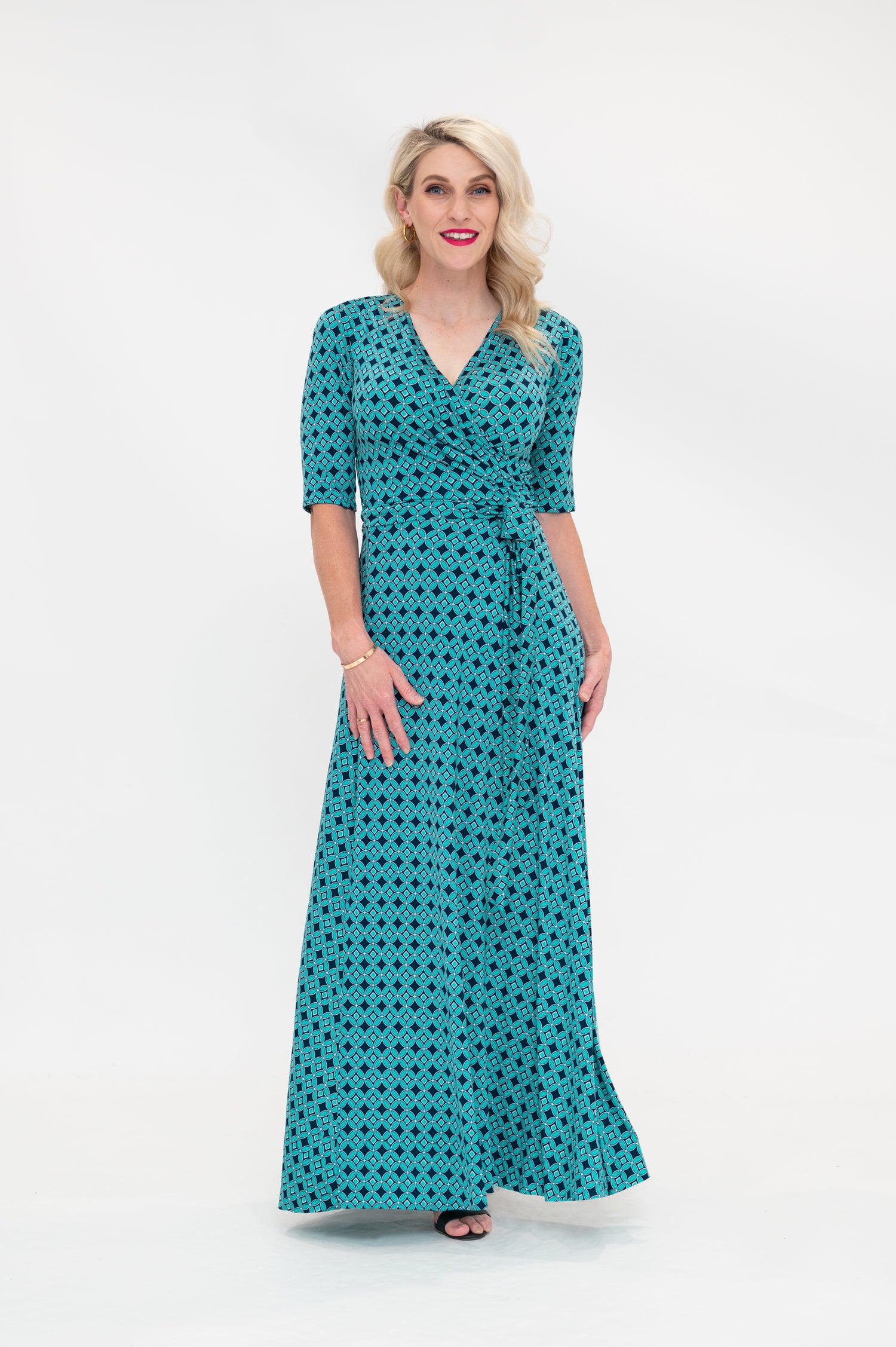 The female model wears a wrap dress in maxi length (floor length) with 3/4 sleeves. The print is a small-scale deep navy, white and teal geometric diamond shape pattern. Front view
