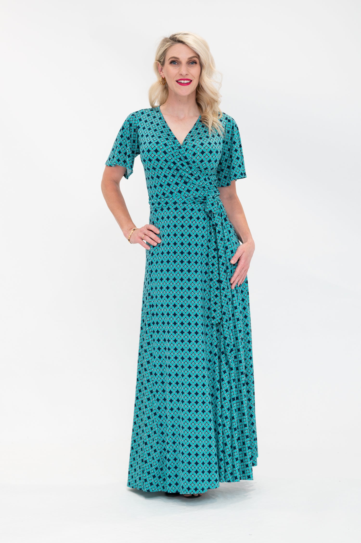 The female model wears a wrap dress in maxi length (floor length) with flutter sleeves. The print is a small-scale deep navy, white and teal geometric diamond shape pattern. Front view