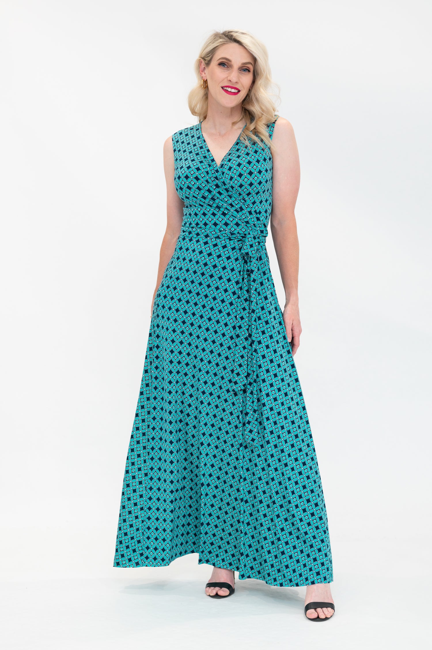 The female model wears a wrap dress in maxi length (floor length) without sleeves. The print is a small-scale deep navy, white and teal geometric diamond shape pattern. Front view