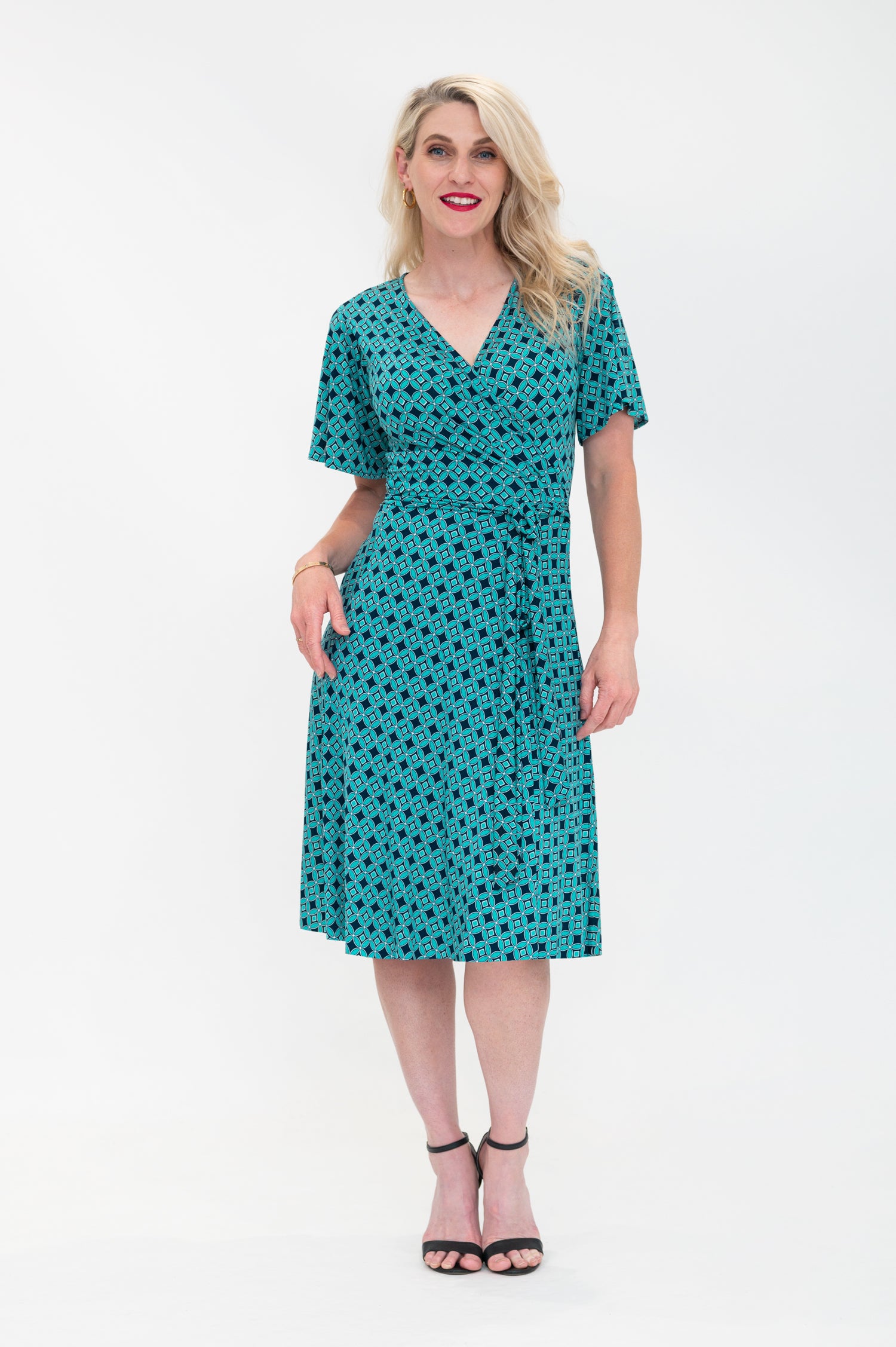 The female model wears a wrap dress in knee length with flutter sleeves. The print is a small-scale deep navy, white and teal geometric diamond shape pattern. Front view