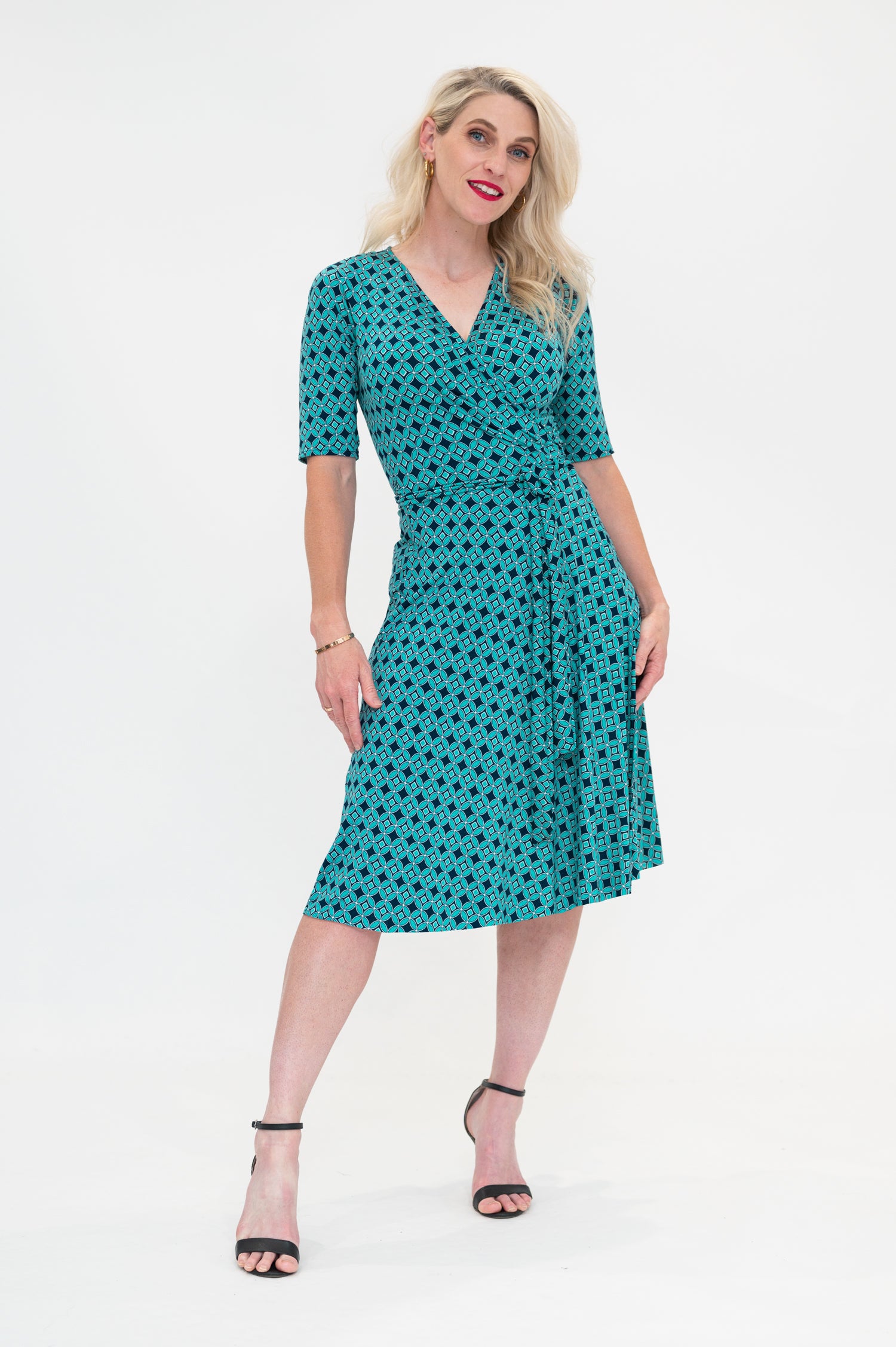 The female model wears a wrap dress in knee length with 3/4 sleeves. The print is a small-scale deep navy, white and teal geometric diamond shape pattern. Front view